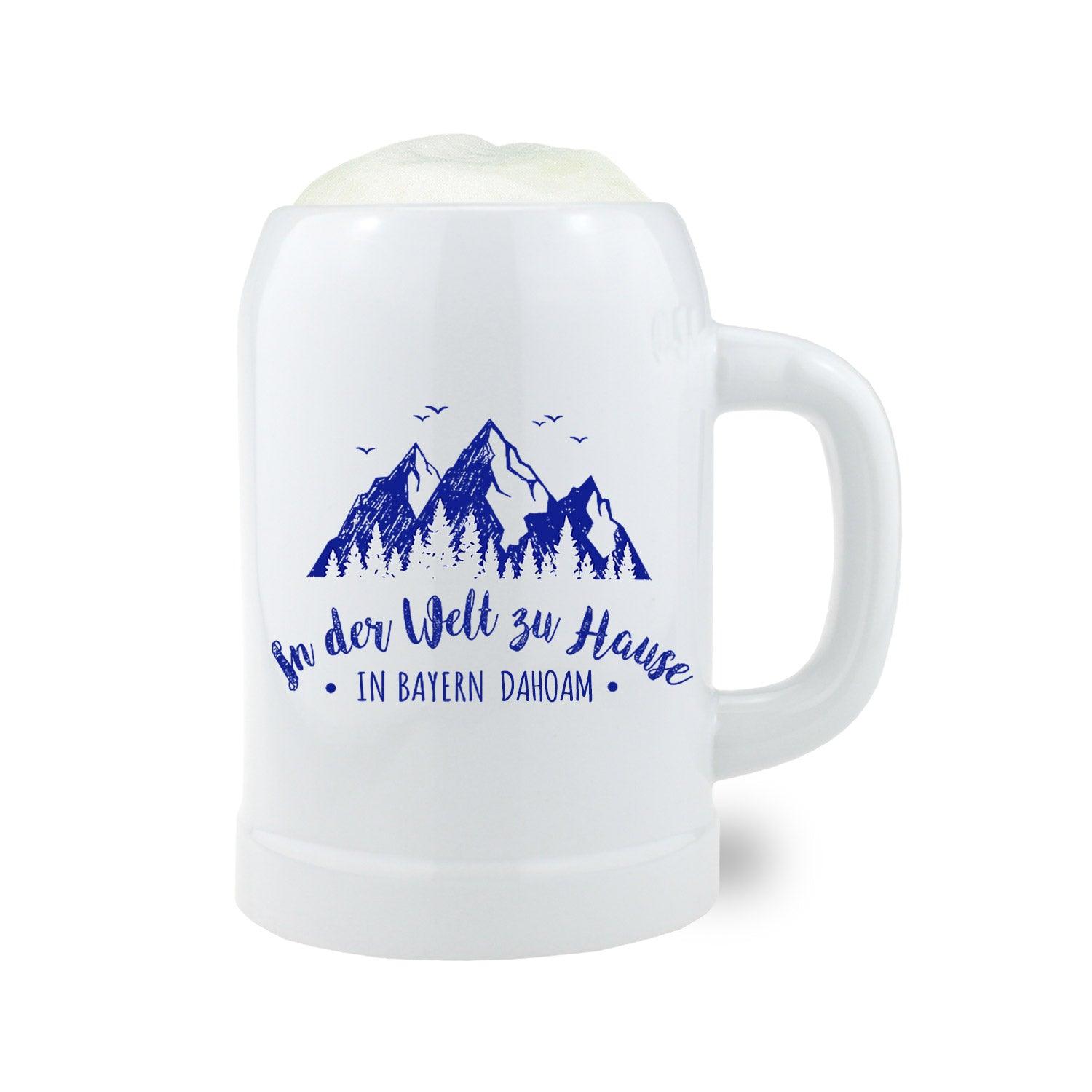 Bierkrug In der Welt zu Hause, in Bayern dahoam mit blauer Alpenlandschaft und bayerischem Spruch auf weißem Porzellan, gefüllt mit Schaum.