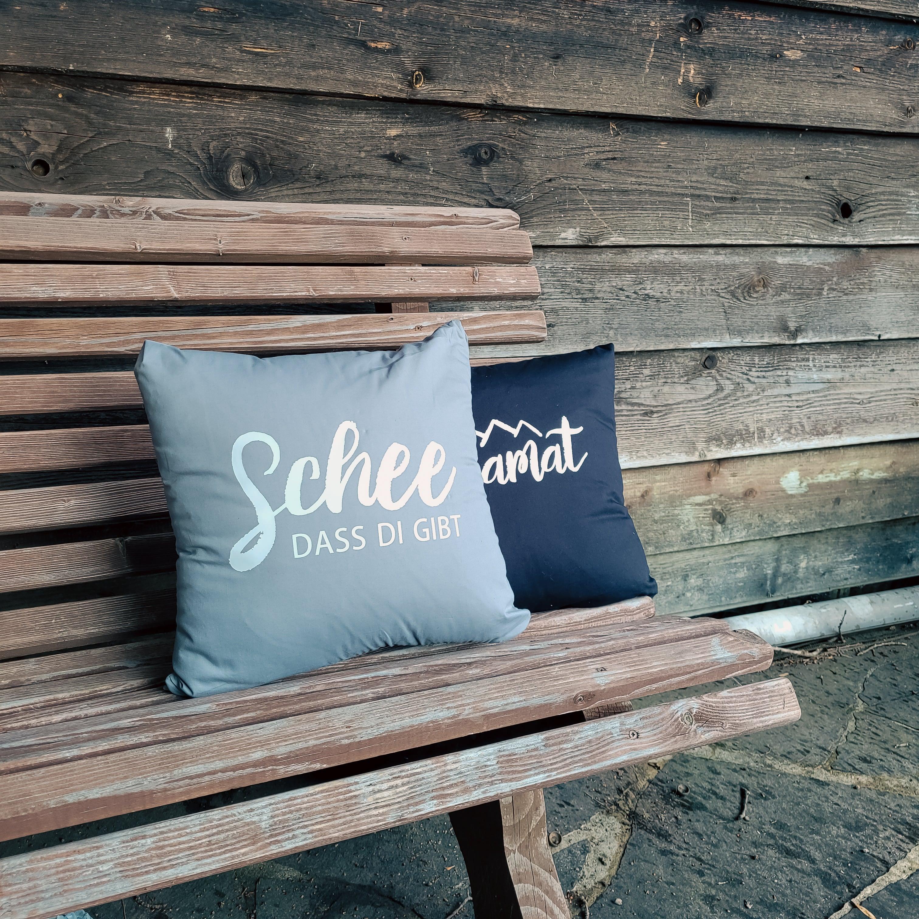 Kissen Schee, dass di gibt auf einer Holzbank, graues Kissen mit weißem Text, dunkles Kissen daneben.