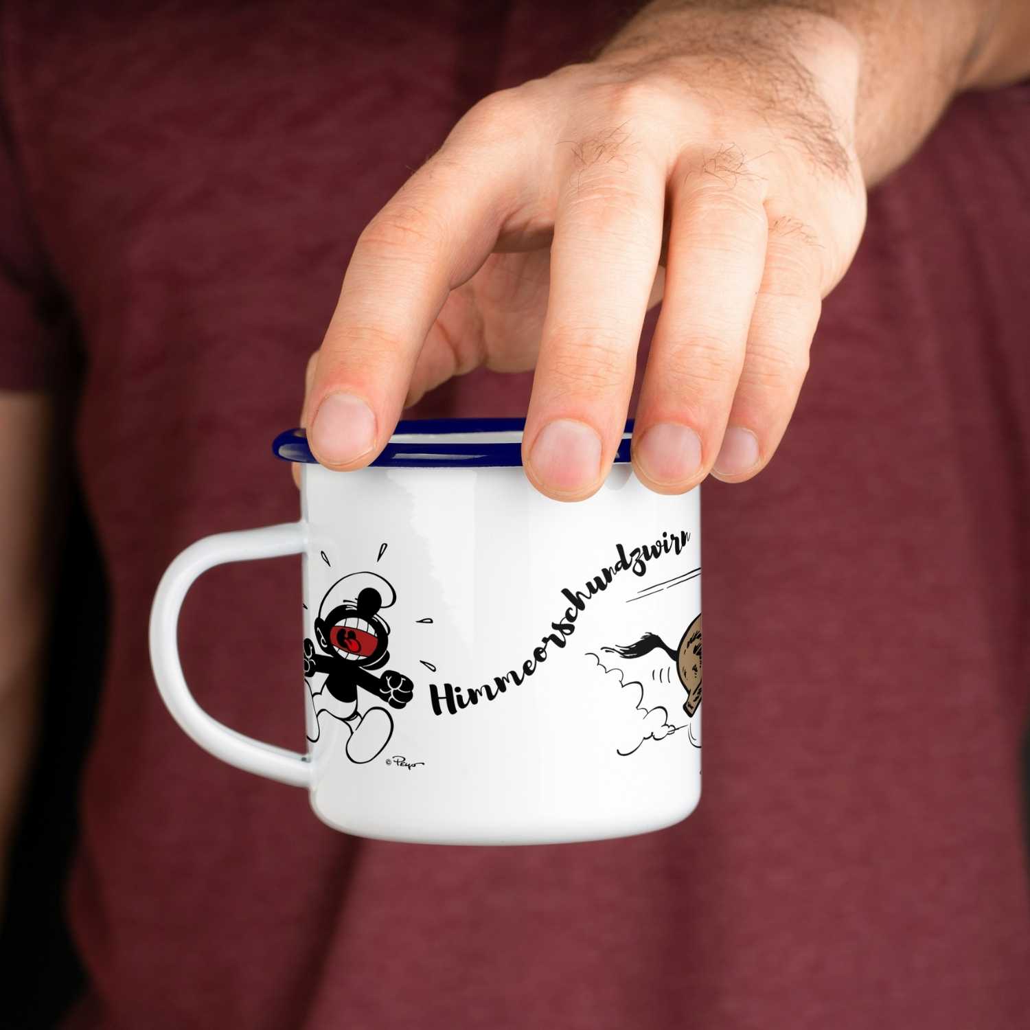 Hand hält ®Die Schlümpfe Emailletasse Himmeorschundzwirn mit lachendem Schlumpf-Motiv und blauem Rand.