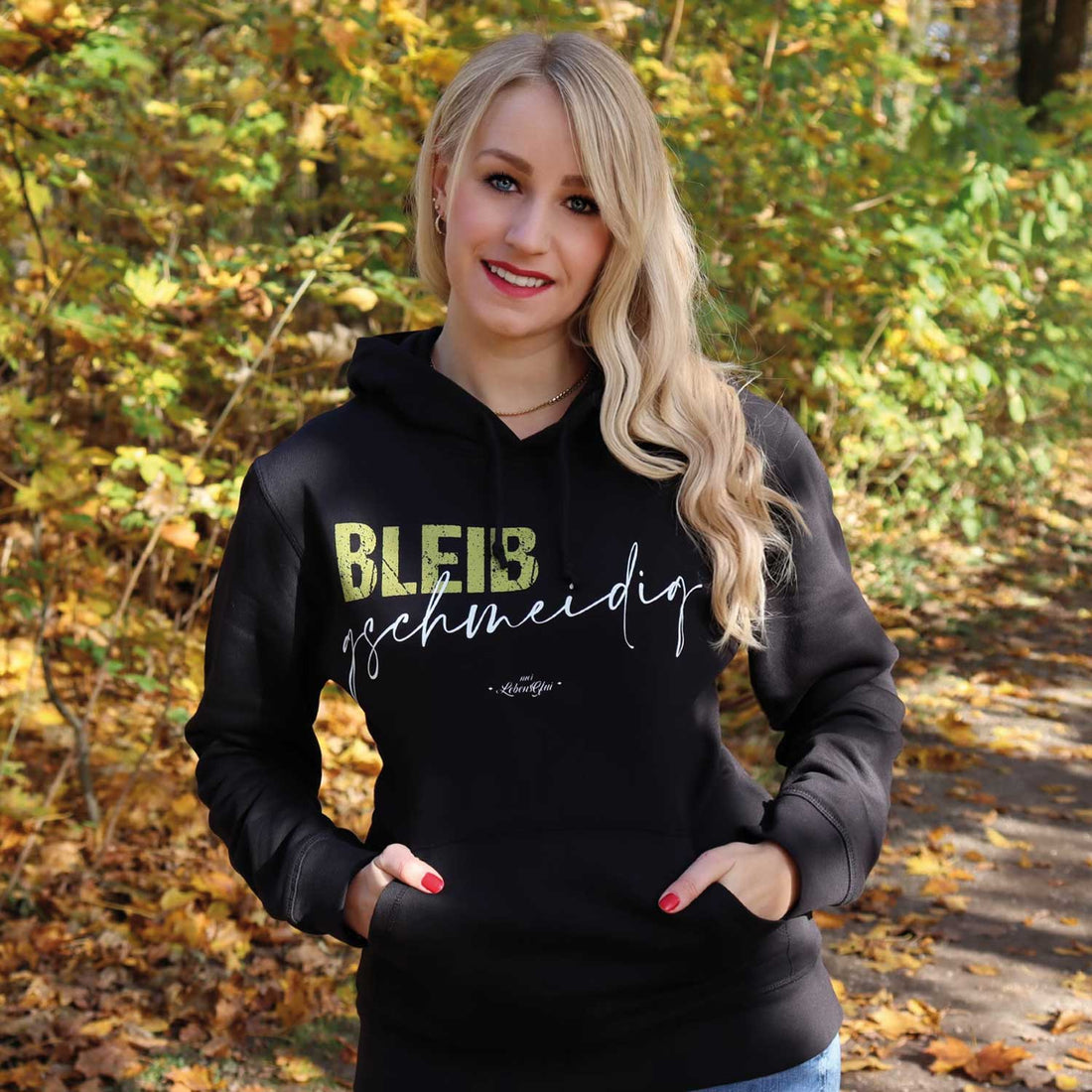 Lächelnde Frau mit blonden Haaren in einem schwarzen Hoodie mit dem Schriftzug "BLEIB geschmeidig" vor herbstlicher Kulisse.