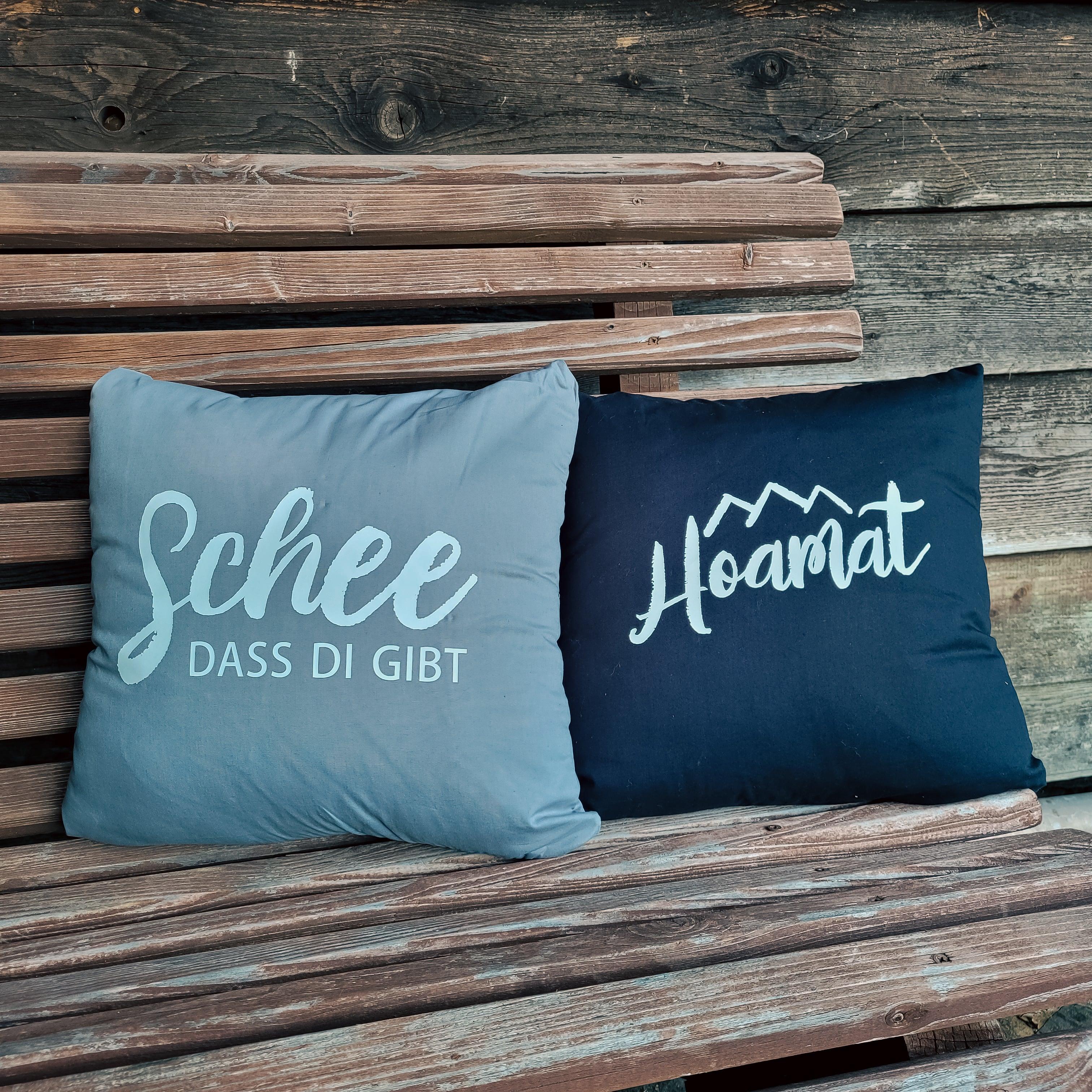 Zwei charmante Kissen "Hoamat" in Grau und Dunkelblau auf einer rustikalen Holzbank.