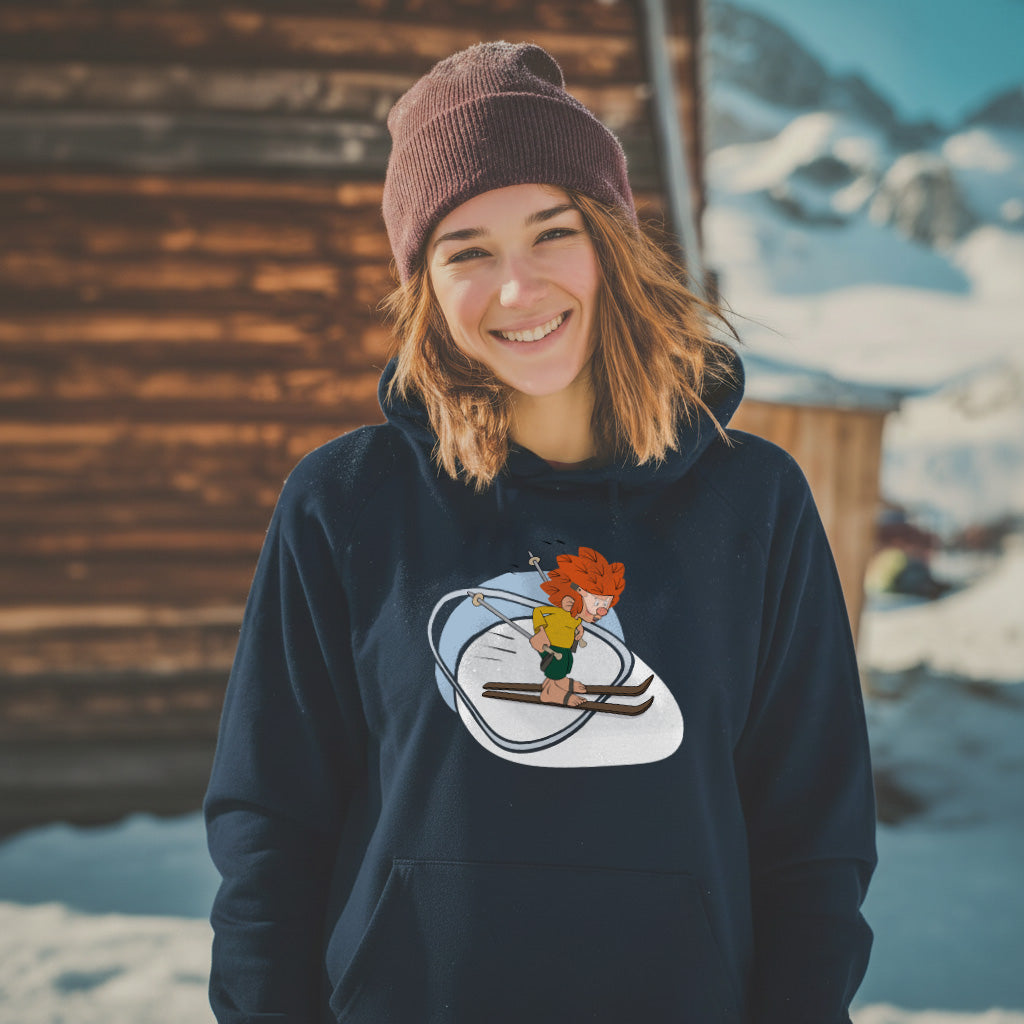 Lachende Frau mit Pumuckl Hoodie Skifahren in Dunkelblau, mit Pumuckl auf Skiern, vor Berghütte im Schnee.