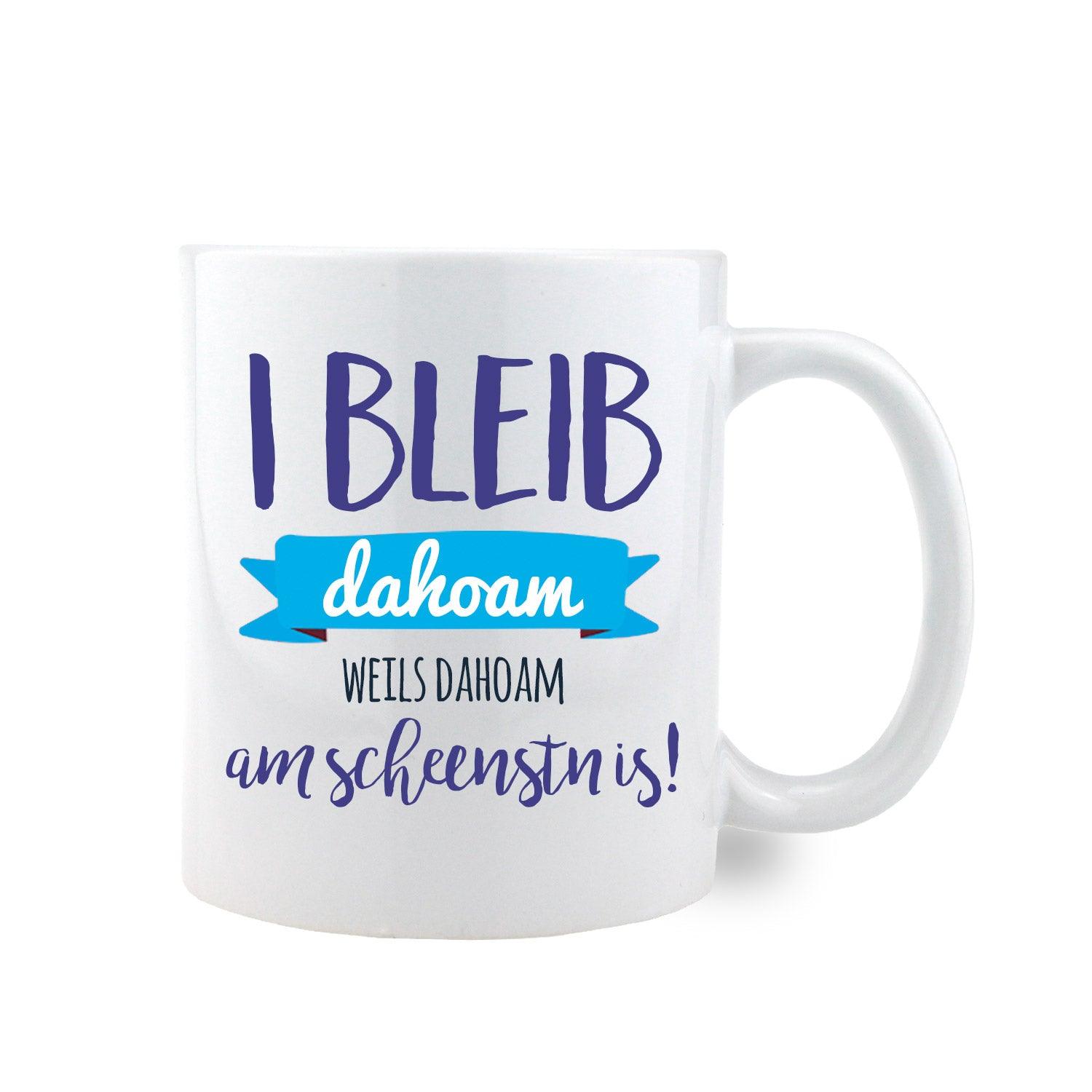 Weiße Keramiktasse mit schwarzem Henkel und Spruch „I bleib dahoam, weils dahoam am scheenstn is!“ gegenüber dem Henkel.