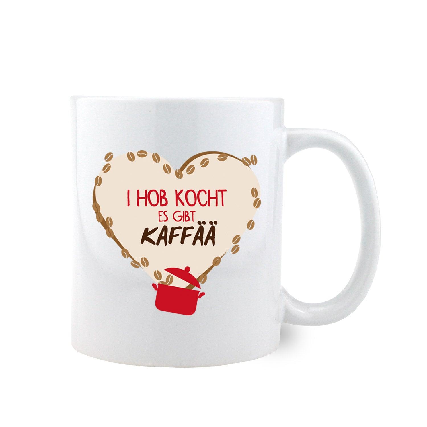 Weiße Keramiktasse mit bauchiger Form, schwarzem Henkel und Aufdruck „I hob kocht – es gibt Kaffää“, ca. 330 ml.