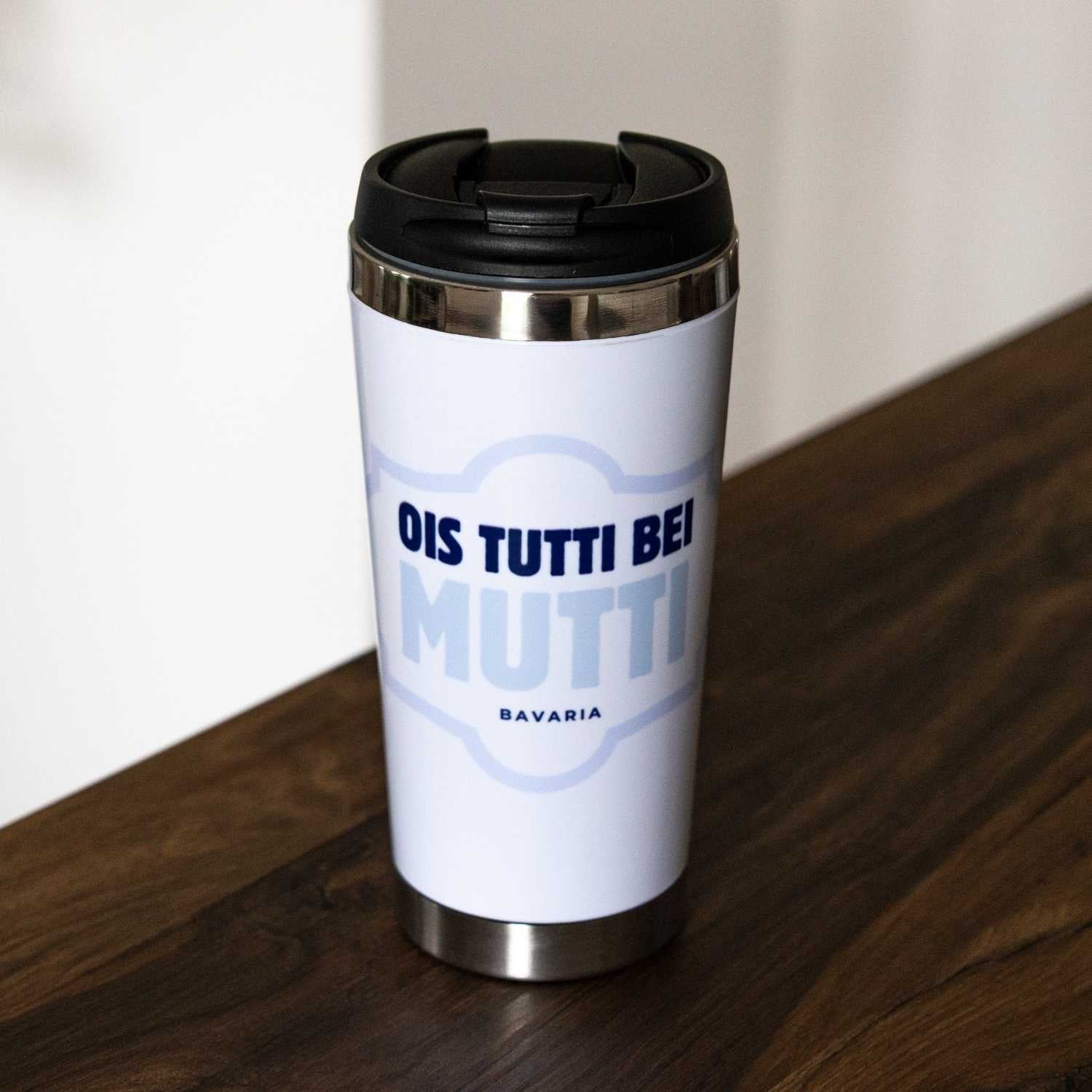 Weißer Thermo-Becher Ois tutti bei Mutti mit blauem Aufdruck und schwarzem Deckel auf Holzoberfläche.