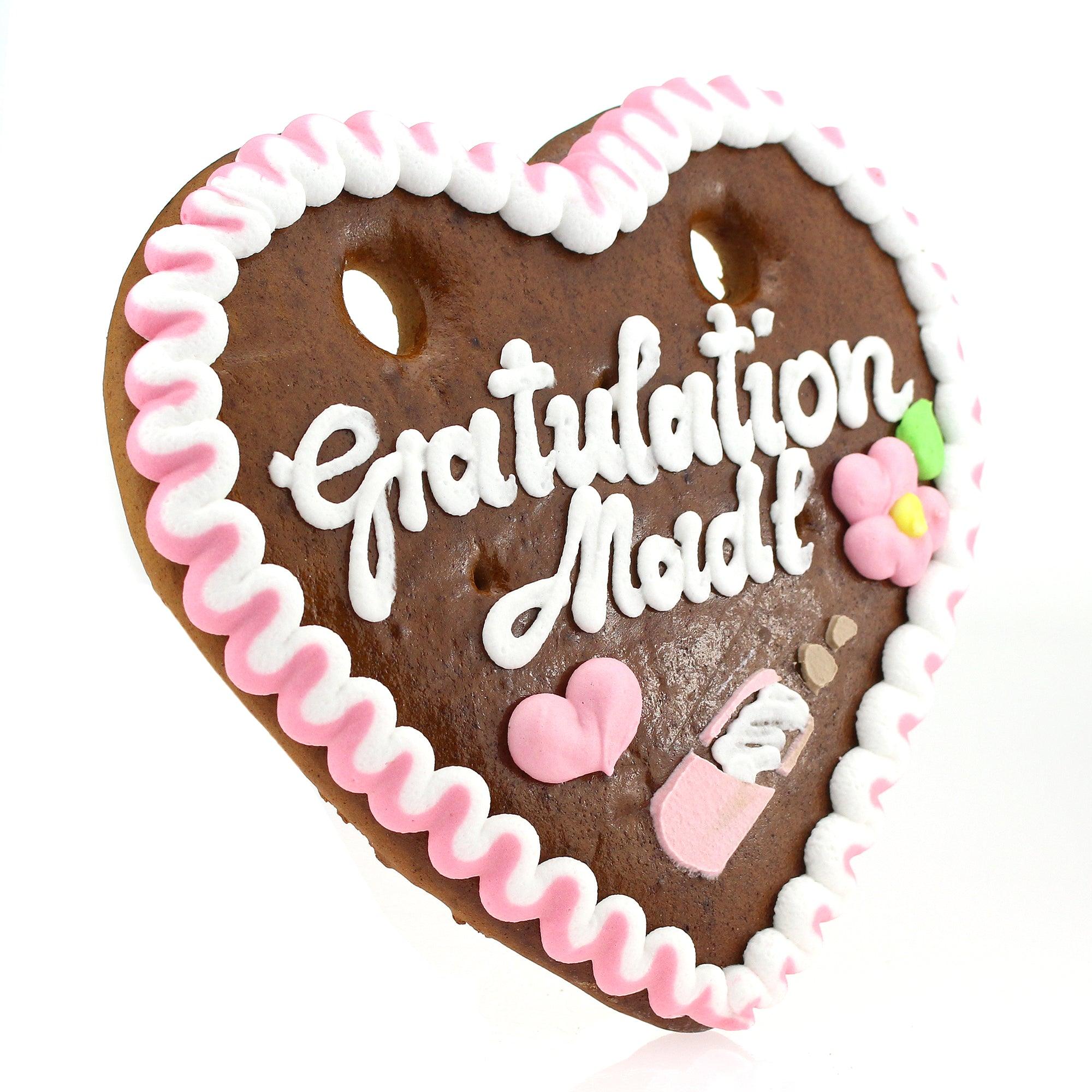 Herzförmiges Lebkuchenherz mit weißer Zuckerschrift „Gratulation Madl“ und rosa Rand aus Zuckerguss.