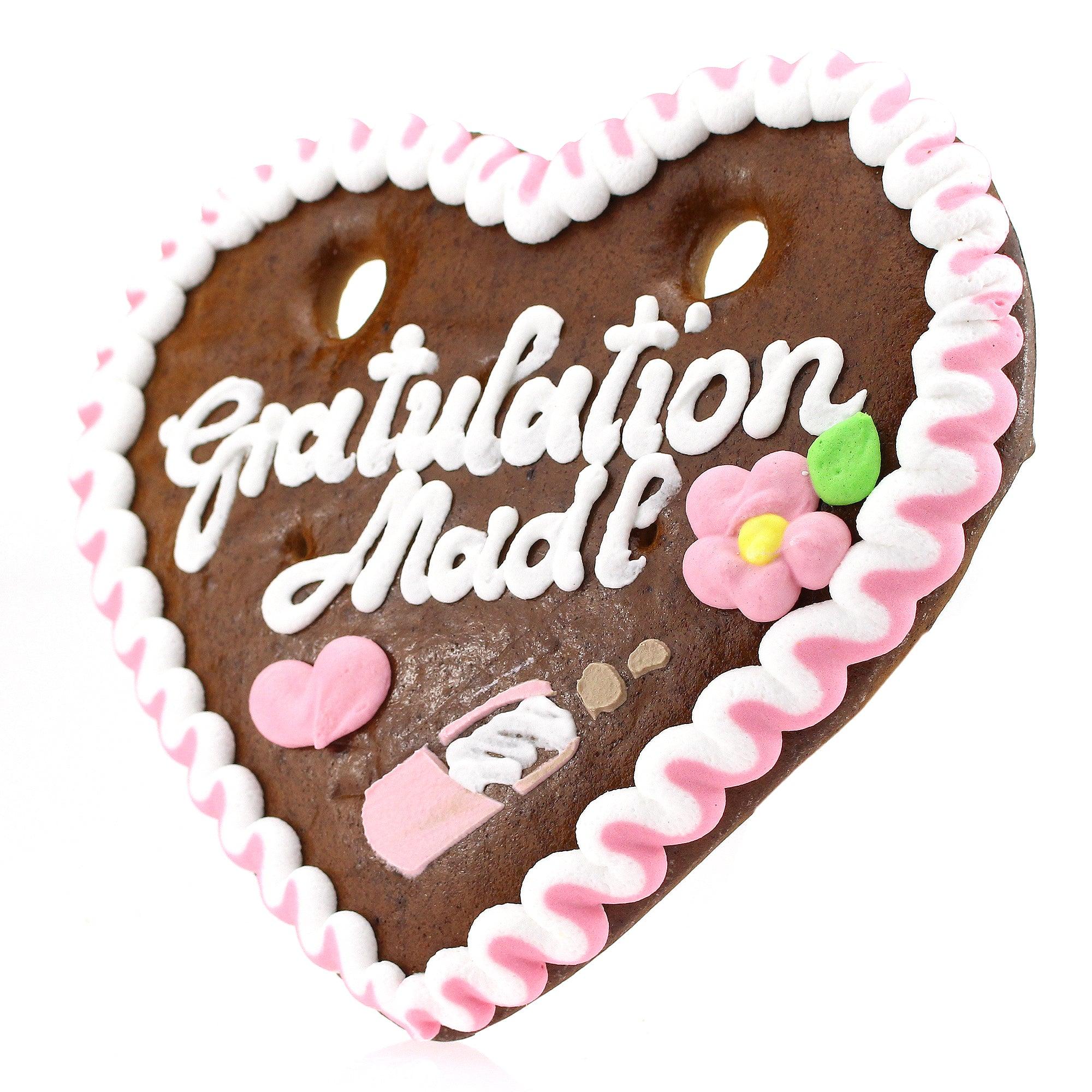 Herzförmiges Lebkuchenherz aus braunem Teig mit rosa Rand, weißer Schrift „Gratulation Madl“, ca. 14 x 17 cm, 100 g.