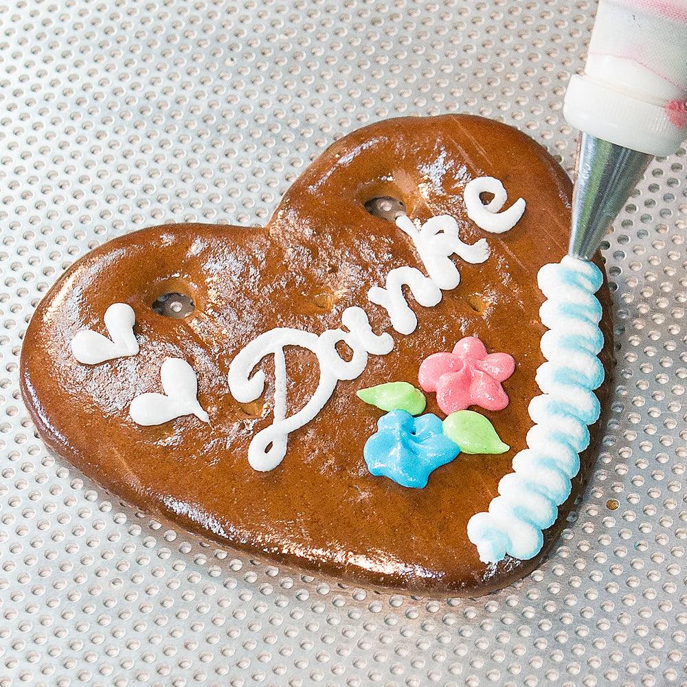 Großes Herz aus braunem Lebkuchen mit weiß-blauem Rand, rosa Blüten und individuellem Wunschtext in Zuckerschrift.