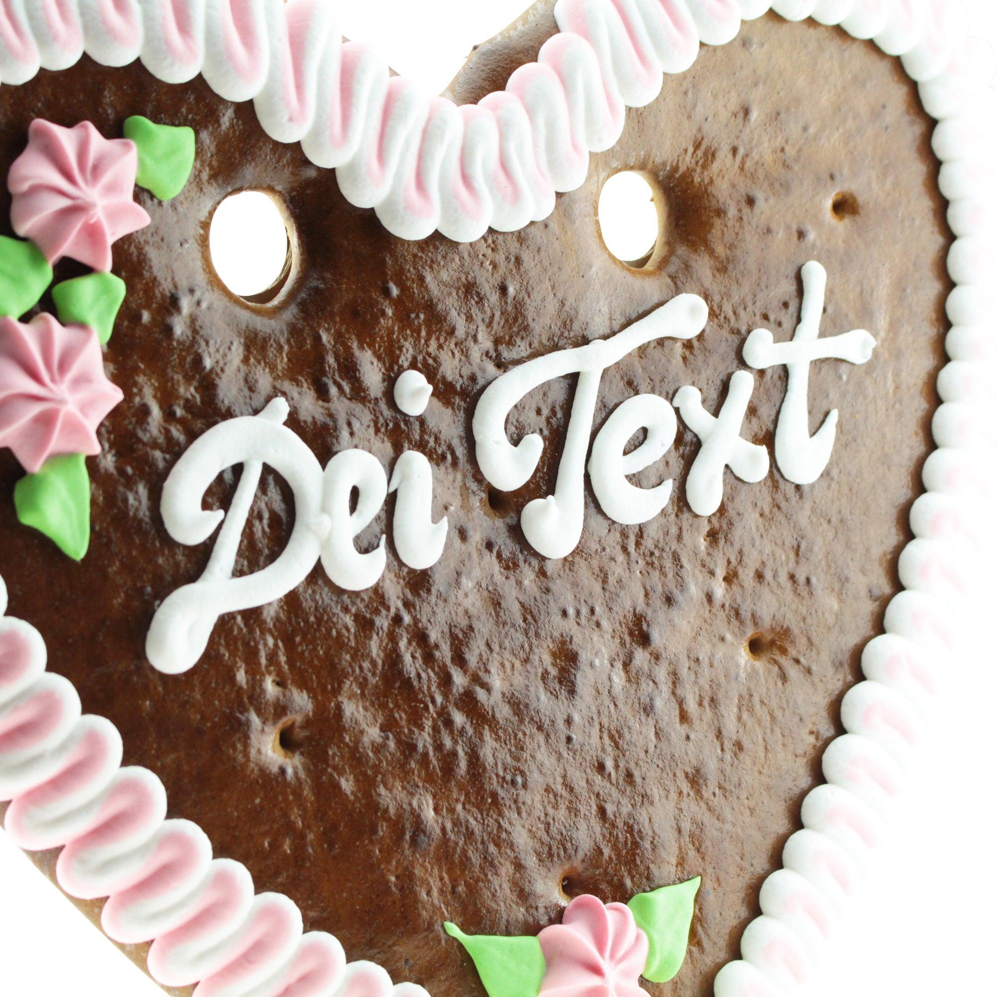Braunes Lebkuchenherz mit weißer Zuckerschrift, rosa Rand, Blumenornamenten und personalisierbarem Text; ca. 32 × 28 cm.