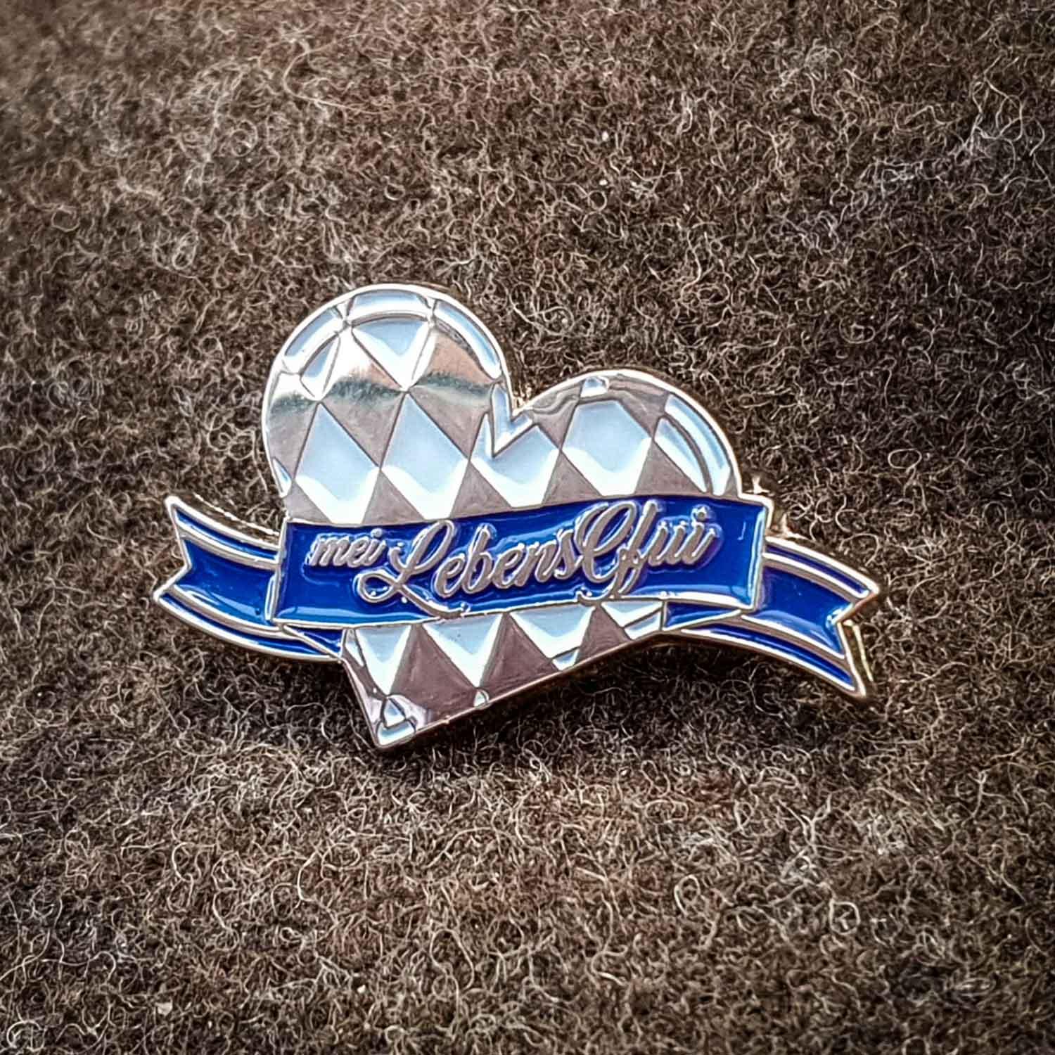 Ovaler Pin mit schwarzem Schriftzug auf silberfarbenem Hintergrund, aus Nickel, ca. 3×2 cm, mit Sicherheitsnadel.