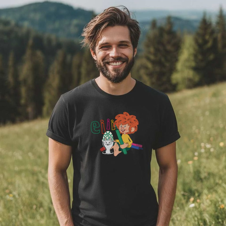 Lachender Mann trägt schwarzes Herren T-Shirt mit Pumuckl und Osterei, naturgeprägte Kulisse.