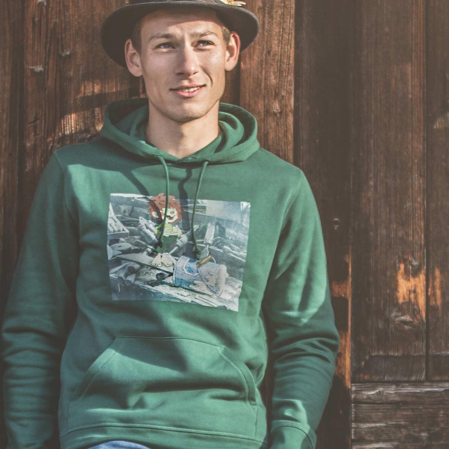 Junger Mann trägt dunkelgrünen Pumuckl Herren Hoodie Werkstatt mit Frontprint, lässig an Holzwand gelehnt.