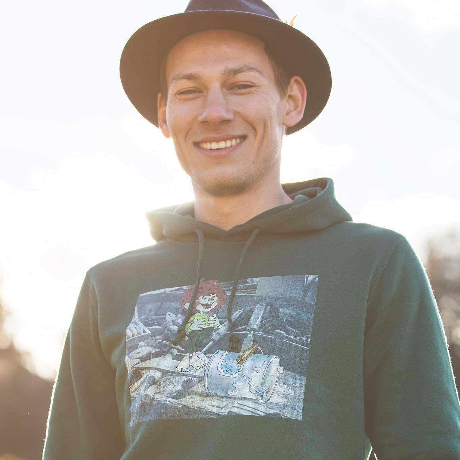 Lächelnder Mann im ®Pumuckl Herren Hoodie Werkstatt, dunkelgrün mit Motiv, bei Sonnenlicht.