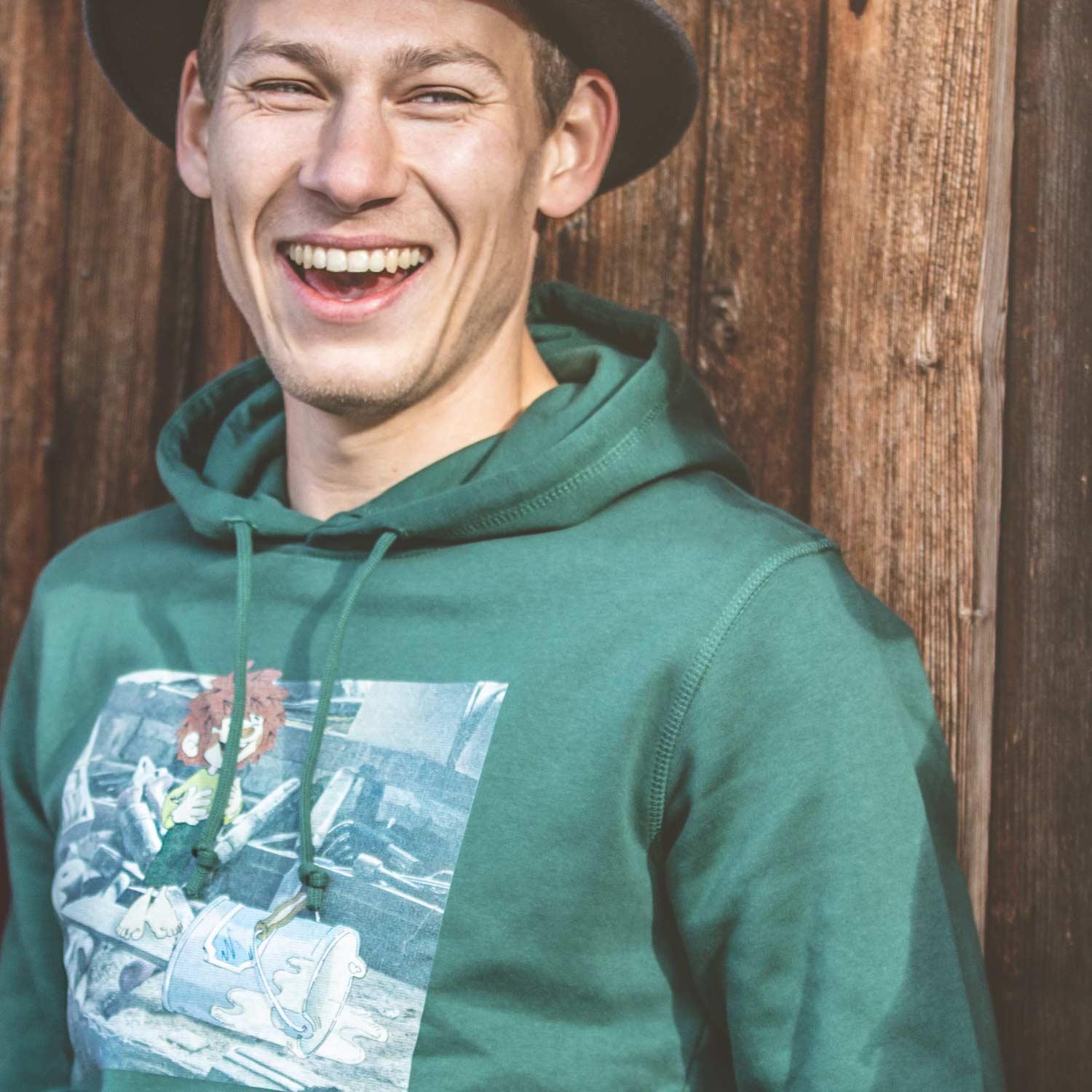 Lächelnder Mann mit ®Pumuckl Herren Hoodie Werkstatt in Grün, der einen Eimer Farbe verschüttet, vor Holzwand.