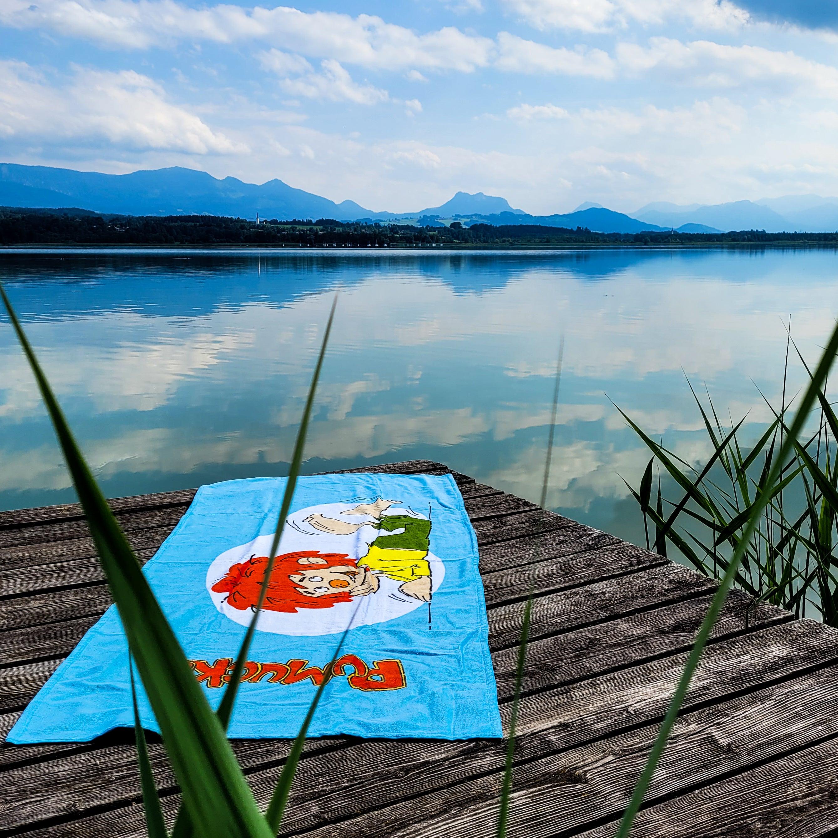 Pumuckl Badetuch XXL auf Holzsteg am See mit Bergpanorama und grünen Schilfhalmen im Vordergrund.