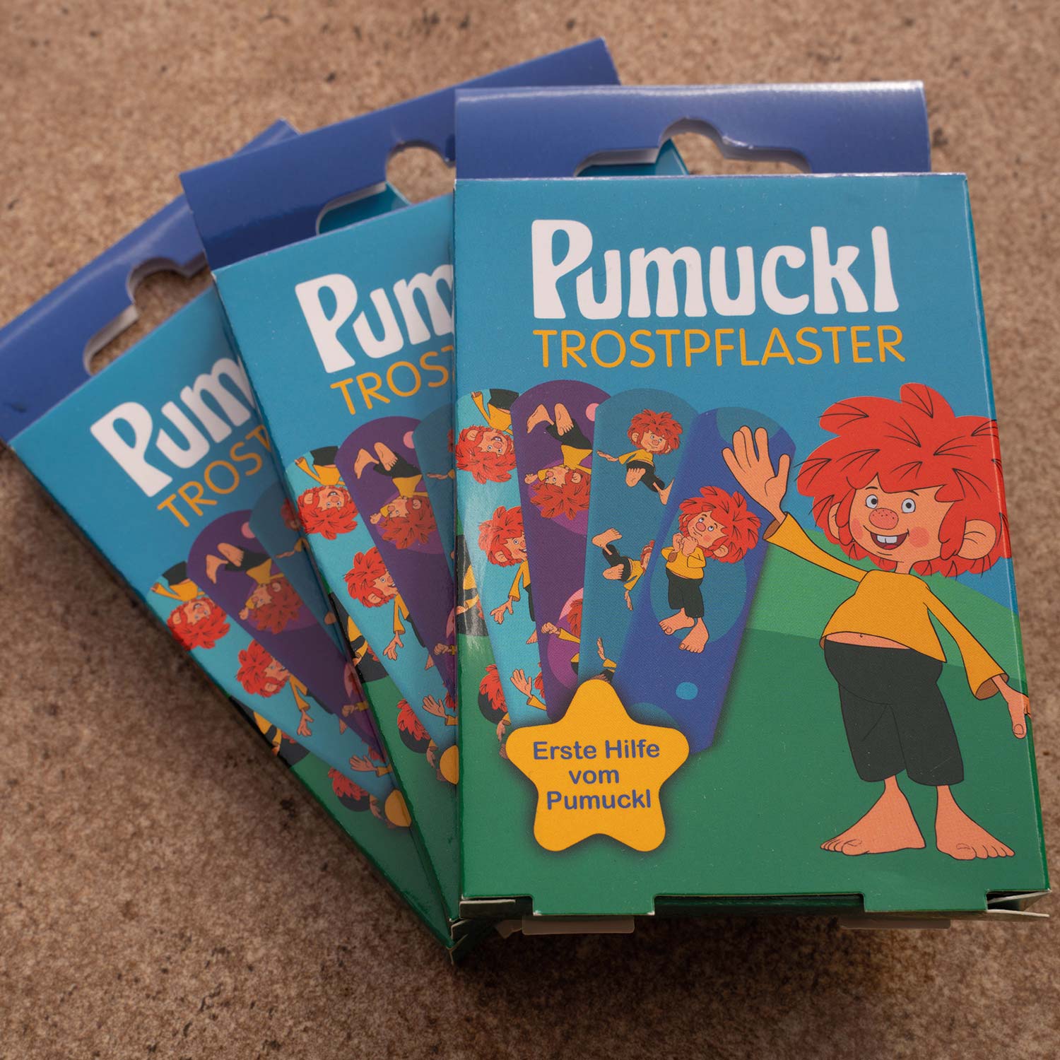 Drei Packungen ®Pumuckl Trost-Pflaster mit bunten Pumuckl-Motiven auf blauem Hintergrund, ideal für Kinder Erste Hilfe.