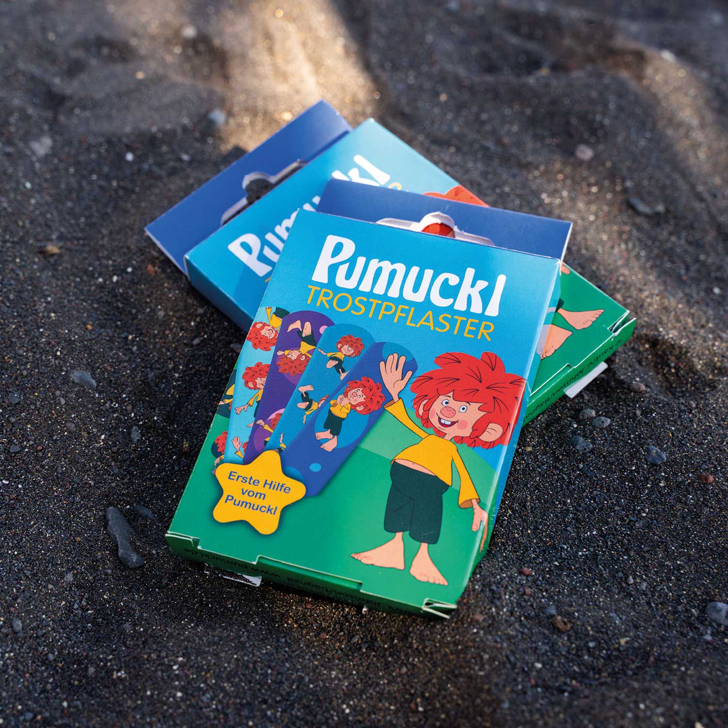 Mehrere Schachteln Pumuckl Trost-Pflaster mit kinderfreundlichem Design, liegend auf schwarzem Sand.