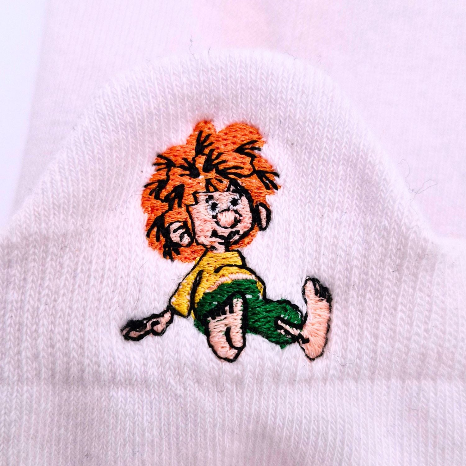 Weißer Sneaker Socke mit gesticktem, fröhlichem Pumuckl, der mit rotem Haar und gelber Kleidung sitzt.