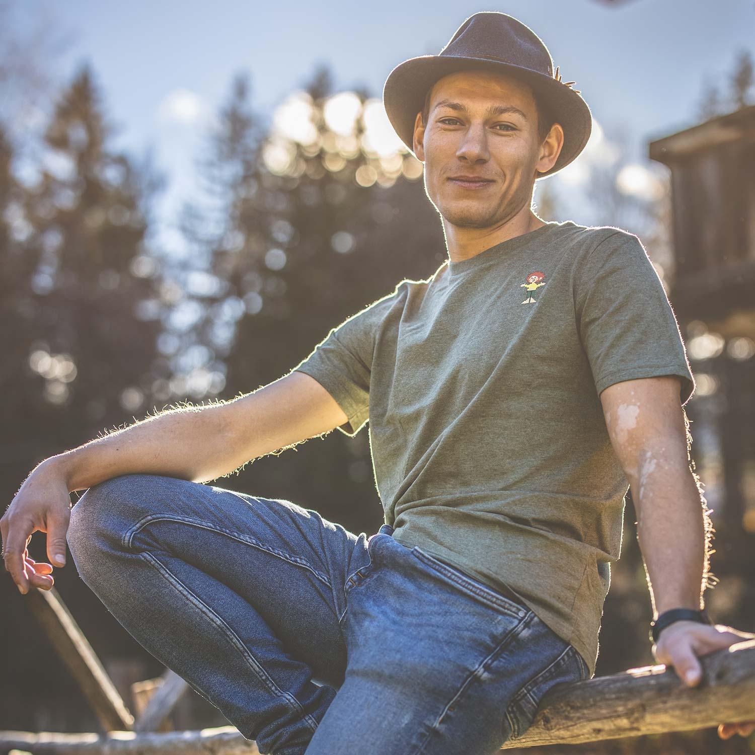 Junger Mann mit Hut trägt grünes ®Pumuckl T-Shirt Pixmuckl auf einer Holzbank in der Natur.