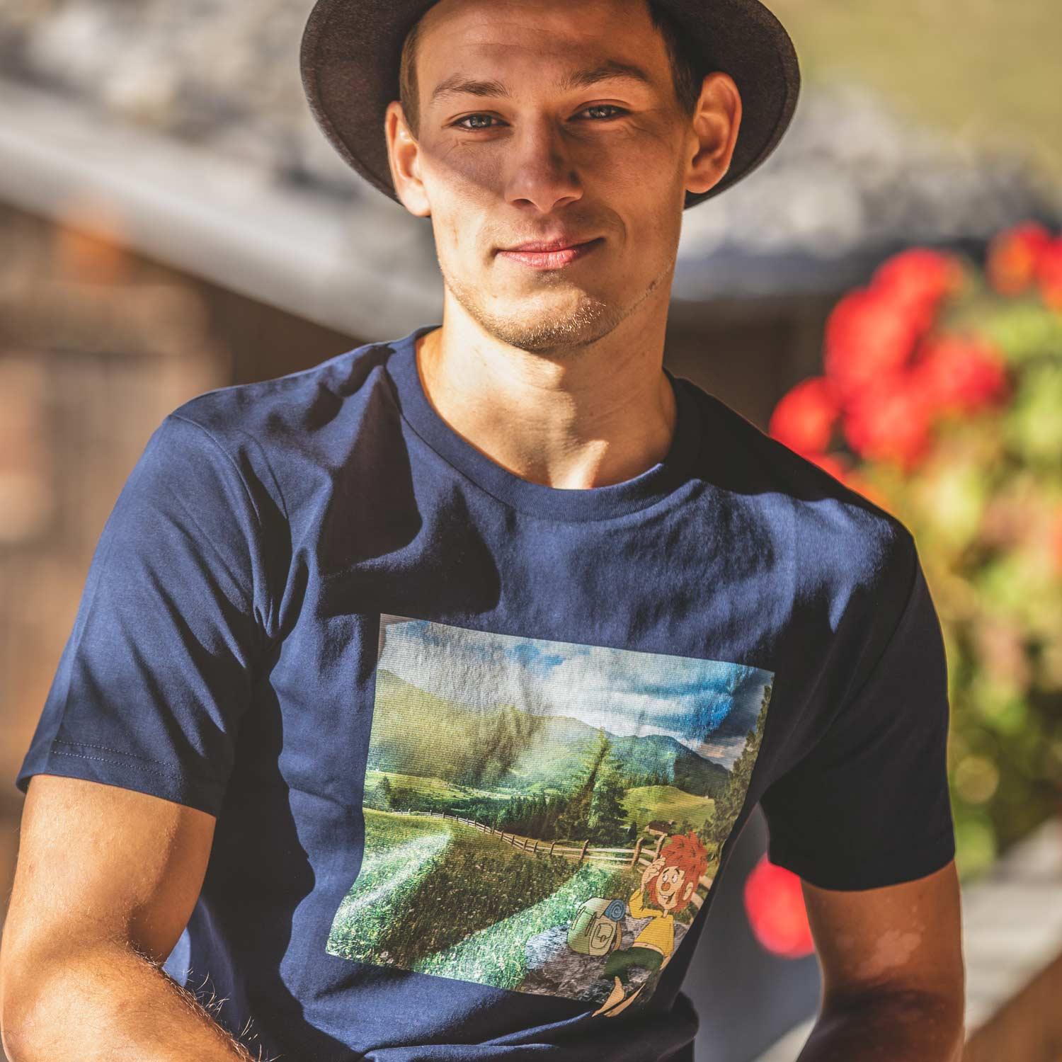 Junger Mann mit dunklem Hut trägt marineblaues ®Pumuckl Herren T-Shirt "Berg-Kobold" mit Berglandschaft und Pumuckl-Motiv.