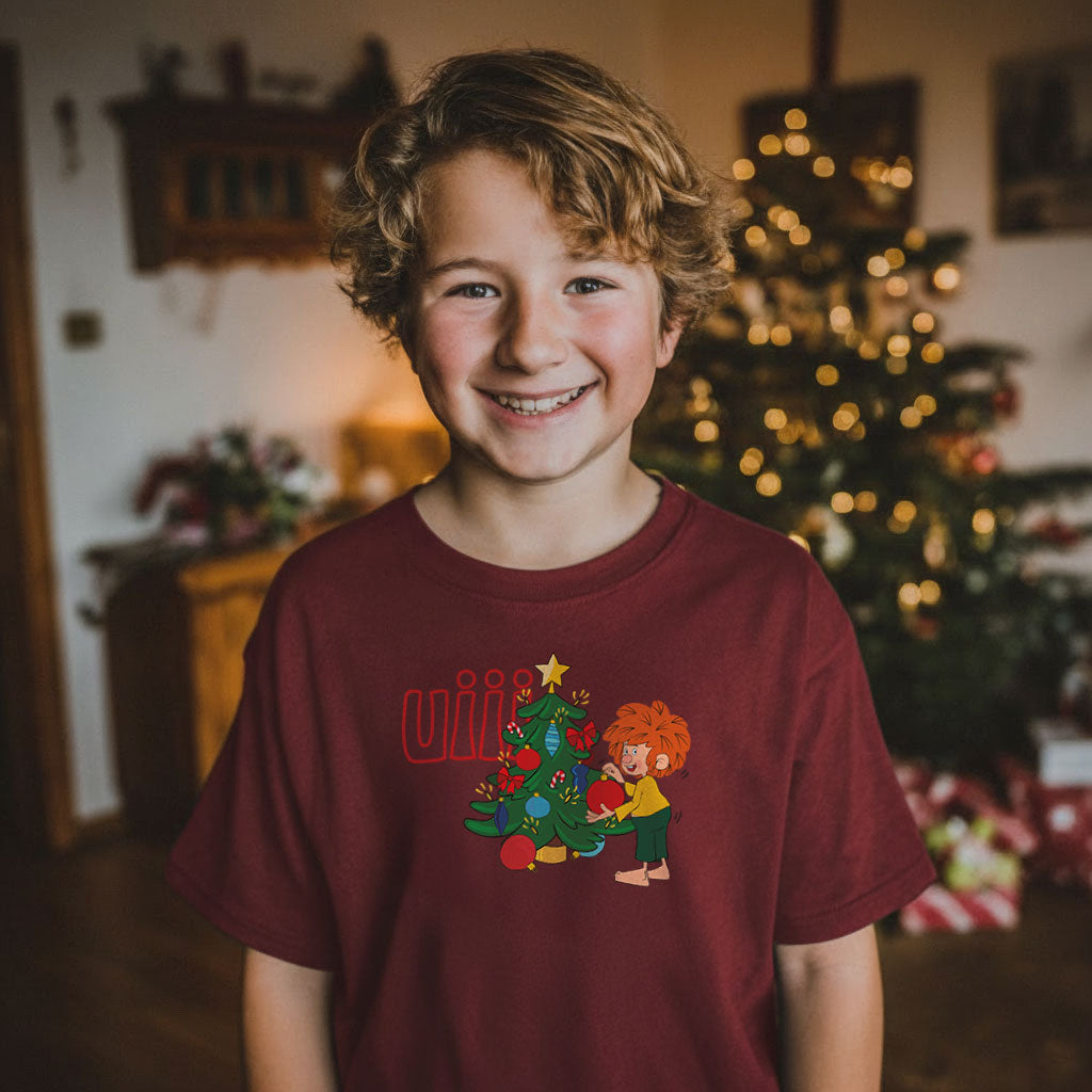Lachender Junge in bordeauxrotem Pumuckl Kinder T-Shirt uiii Weihnachten mit geschmücktem Tannenbaum im Hintergrund.