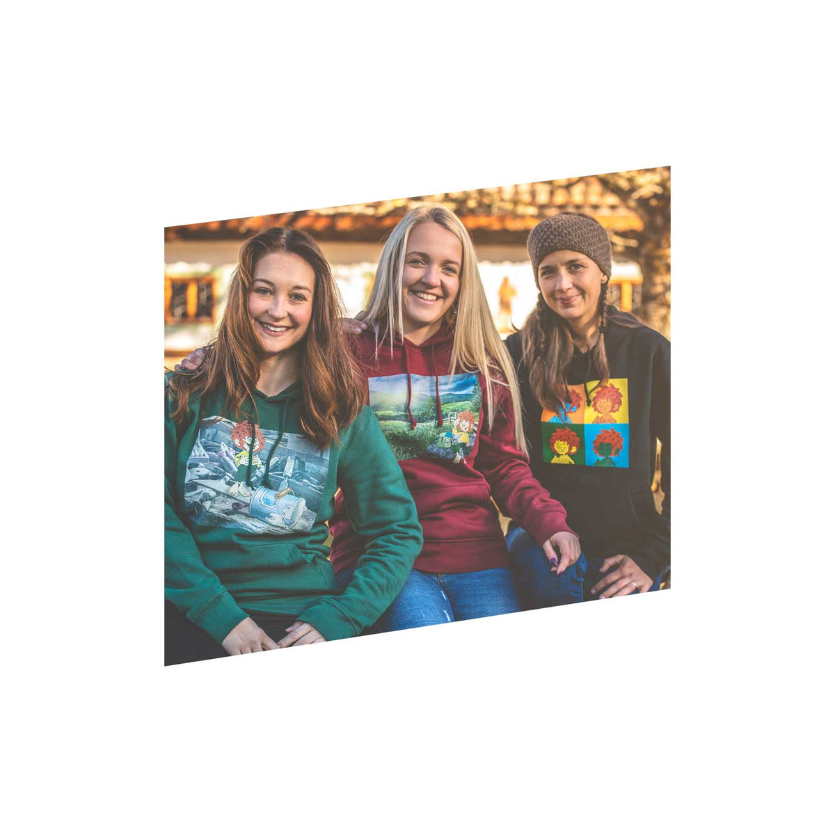 Drei junge Frauen tragen bunte Hoodies mit comicähnlichen Motiven und lächeln in die Kamera.