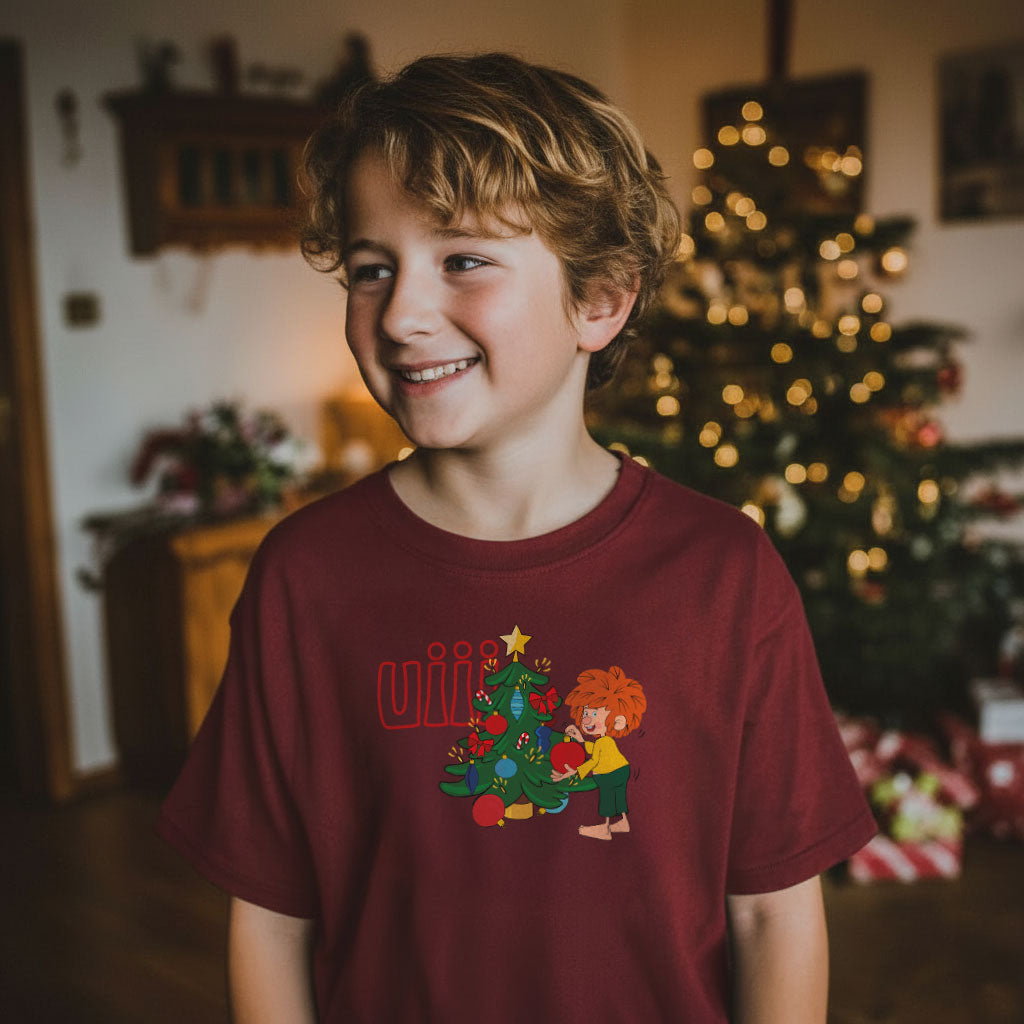 Lächelnder Junge in bordeauxrotem Pumuckl Kinder T-Shirt mit uiih Weihnachten Motiv und Christbaum im Hintergrund.