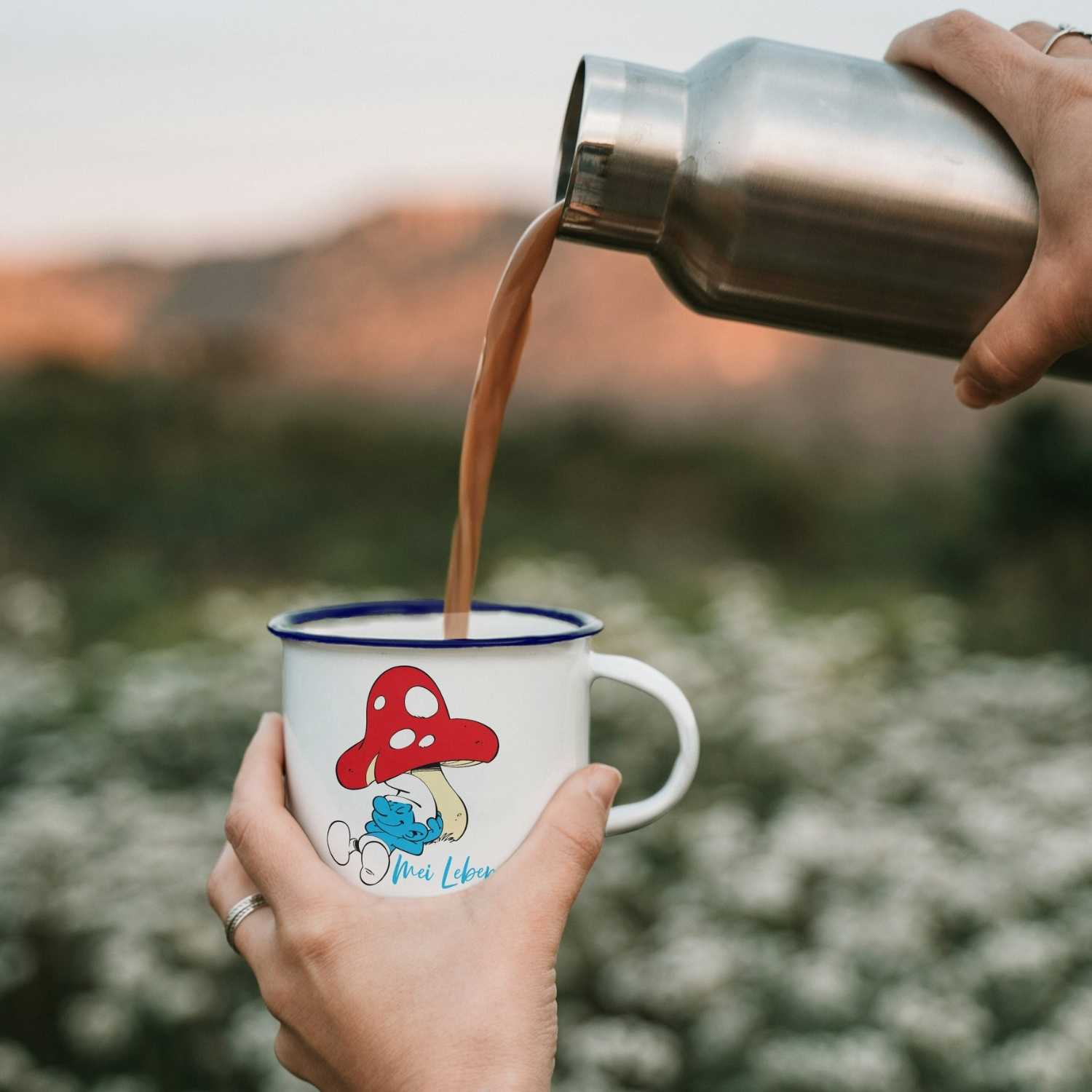 ®Die Schlümpfe Emailletasse Do legst di nieda mit Pilz-Motiv, befüllt mit Kaffee aus Thermosflasche, in Hand gehalten