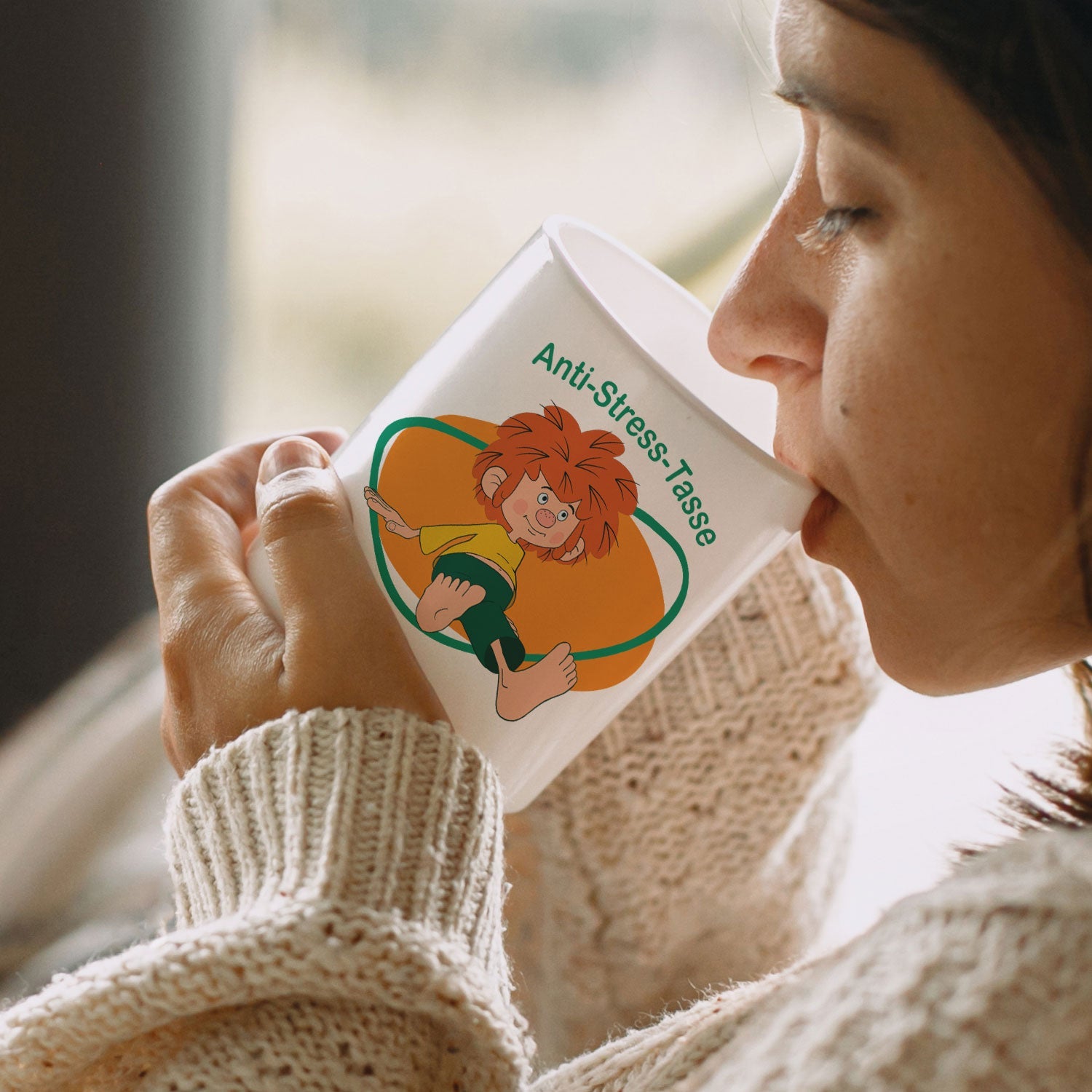 Frau trinkt aus ®Pumuckl Anti-Stress-Tasse mit Kobold-Motiv, kuscheligem Pullover in gemütlicher Atmosphäre.